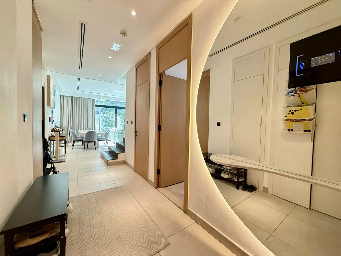 Hallway with beige tile floor, light wood doors, and a large, illuminated, curved mirror reflecting a shoe rack and TV. Living room visible in background.