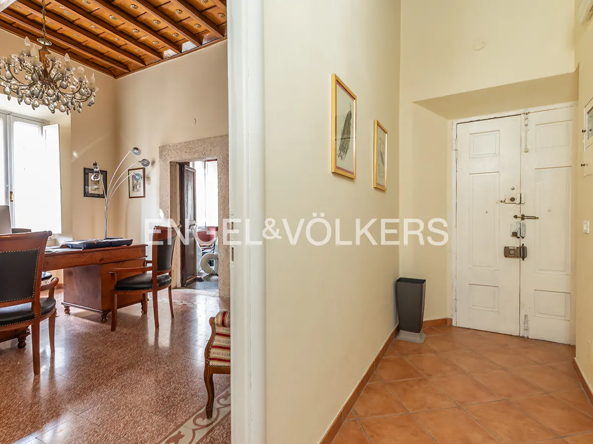 Interior view of a hallway with a white door and a glimpse into an office with a wooden desk and chandelier.