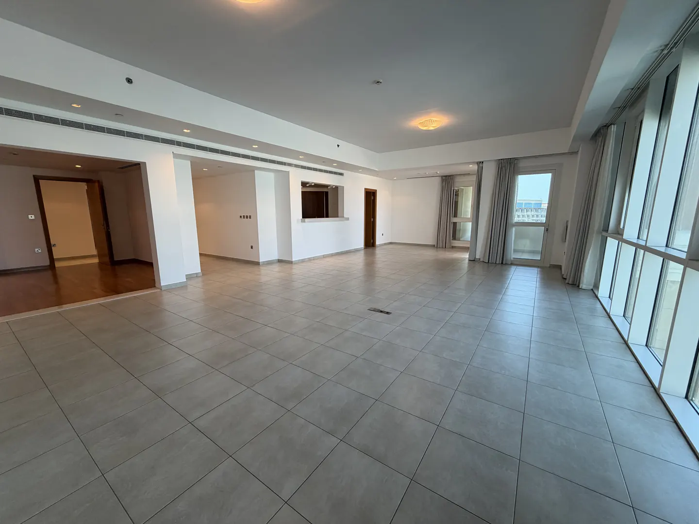 Spacious, empty room with light grey tiled floors, white walls, and floor-to-ceiling windows with curtains.