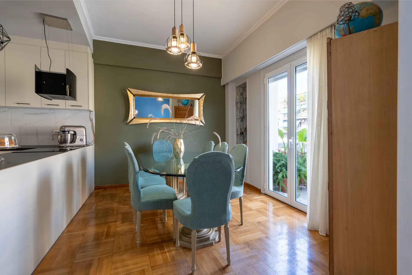 A dining area with a glass table, blue chairs, and a gold-framed mirror on an olive green wall. A globe sits atop a wooden cabinet.