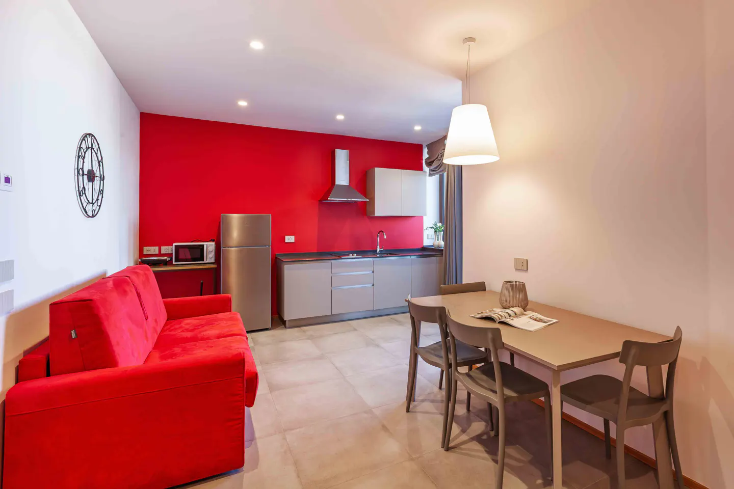Bright apartment interior with a red accent wall, stainless steel appliances, and a red sofa. A dining table with four chairs sits nearby.