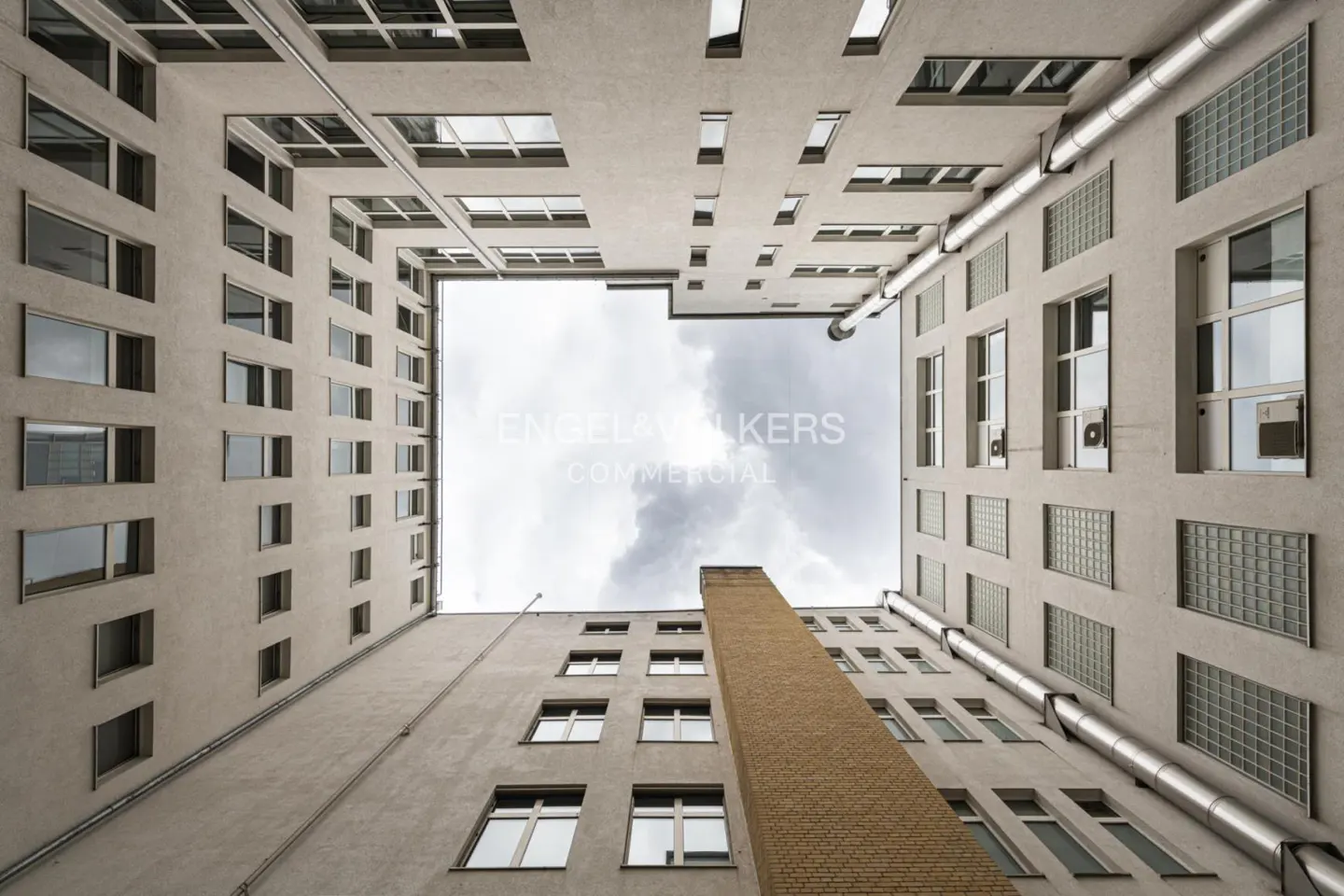 Looking up at a square courtyard of a tall, light gray building with many windows and a brick chimney against a cloudy sky.