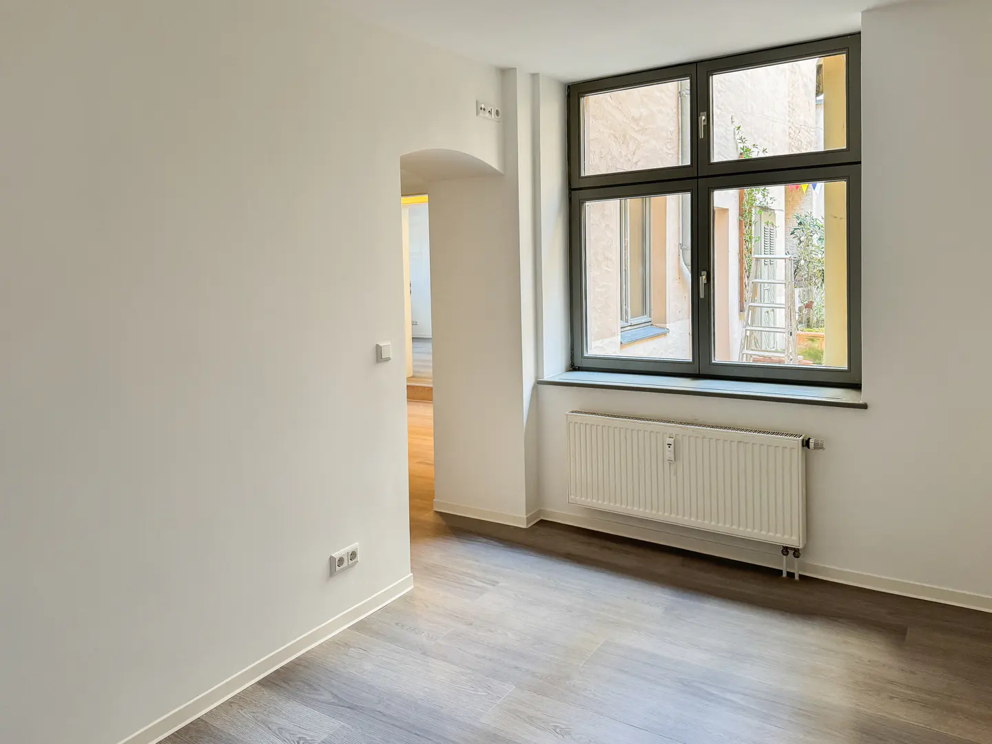 Empty room with white walls, gray wood floors, and a dark-framed window showing a courtyard with a ladder. A white radiator is under the window.