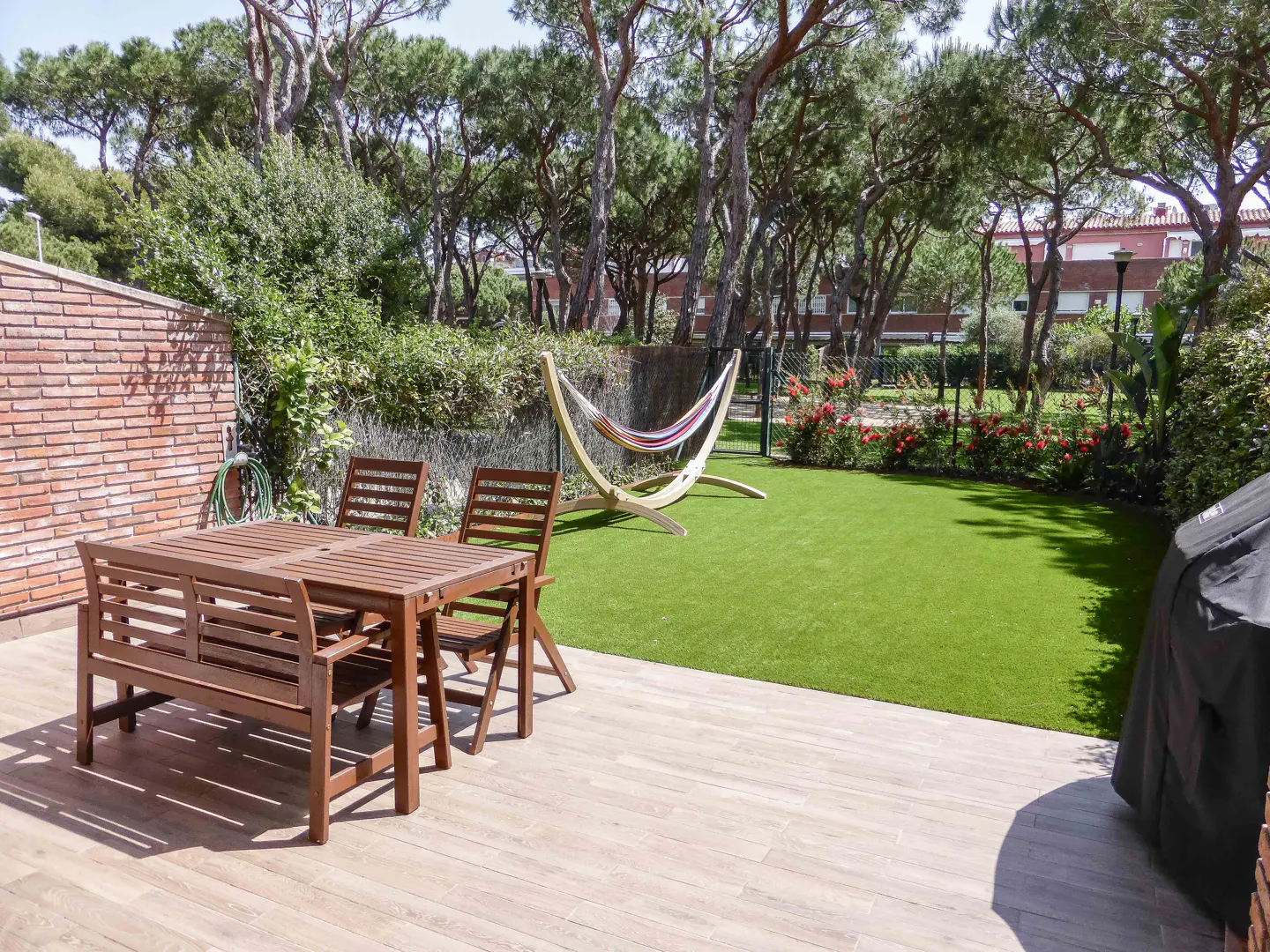 Outdoor patio with wood table, chairs, and bench on a tiled patio. A hammock sits on green grass surrounded by trees and flowers.