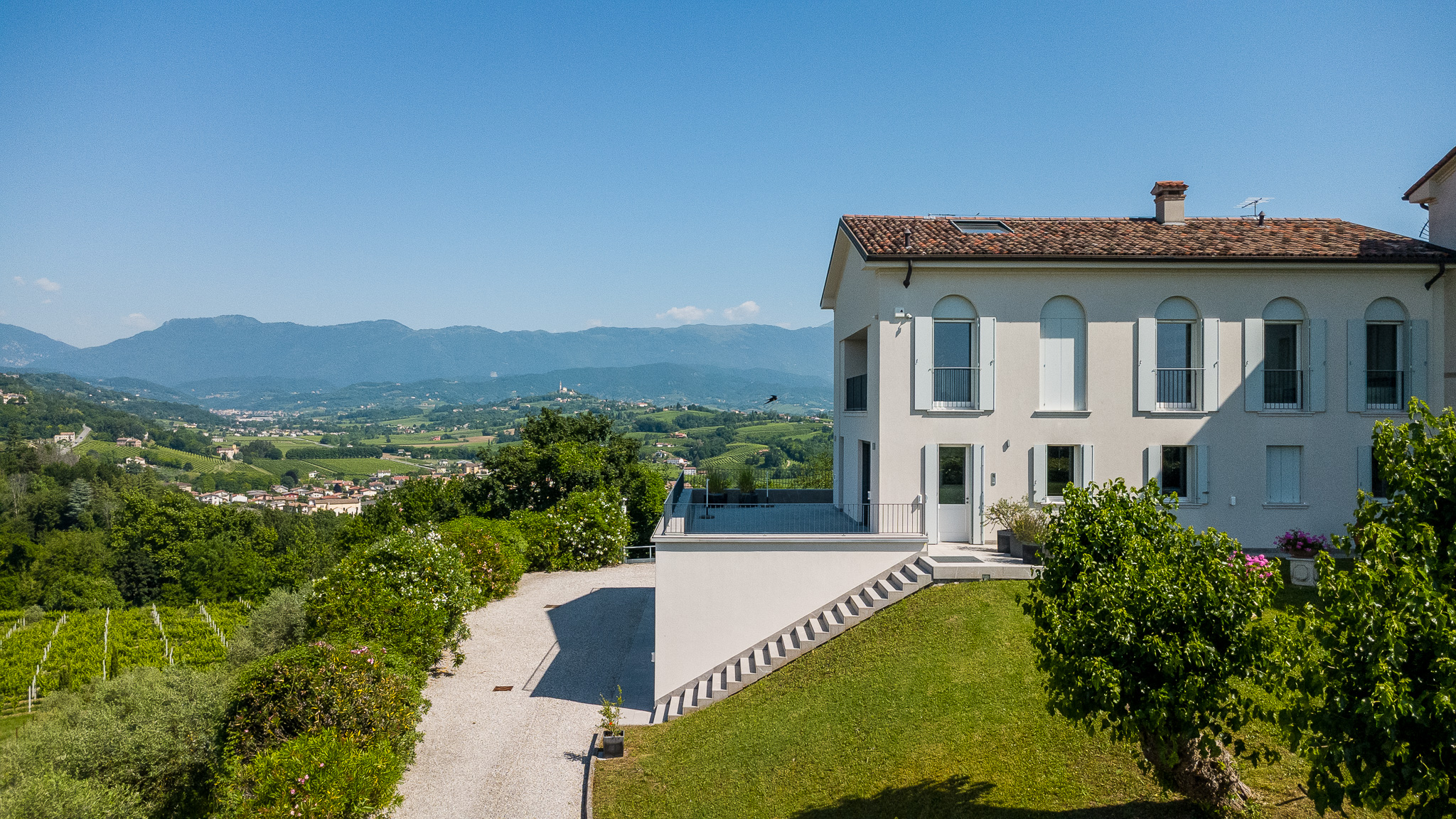 A cream-colored villa with a tiled roof overlooks a lush green valley and distant mountains on a sunny day.