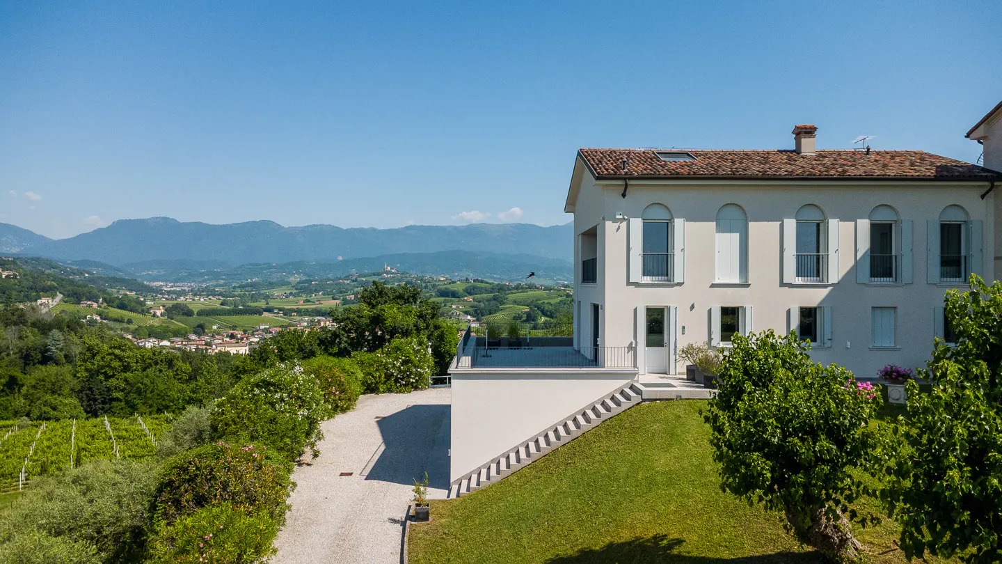 A cream-colored villa with a tiled roof overlooks a lush green valley and distant mountains on a sunny day.