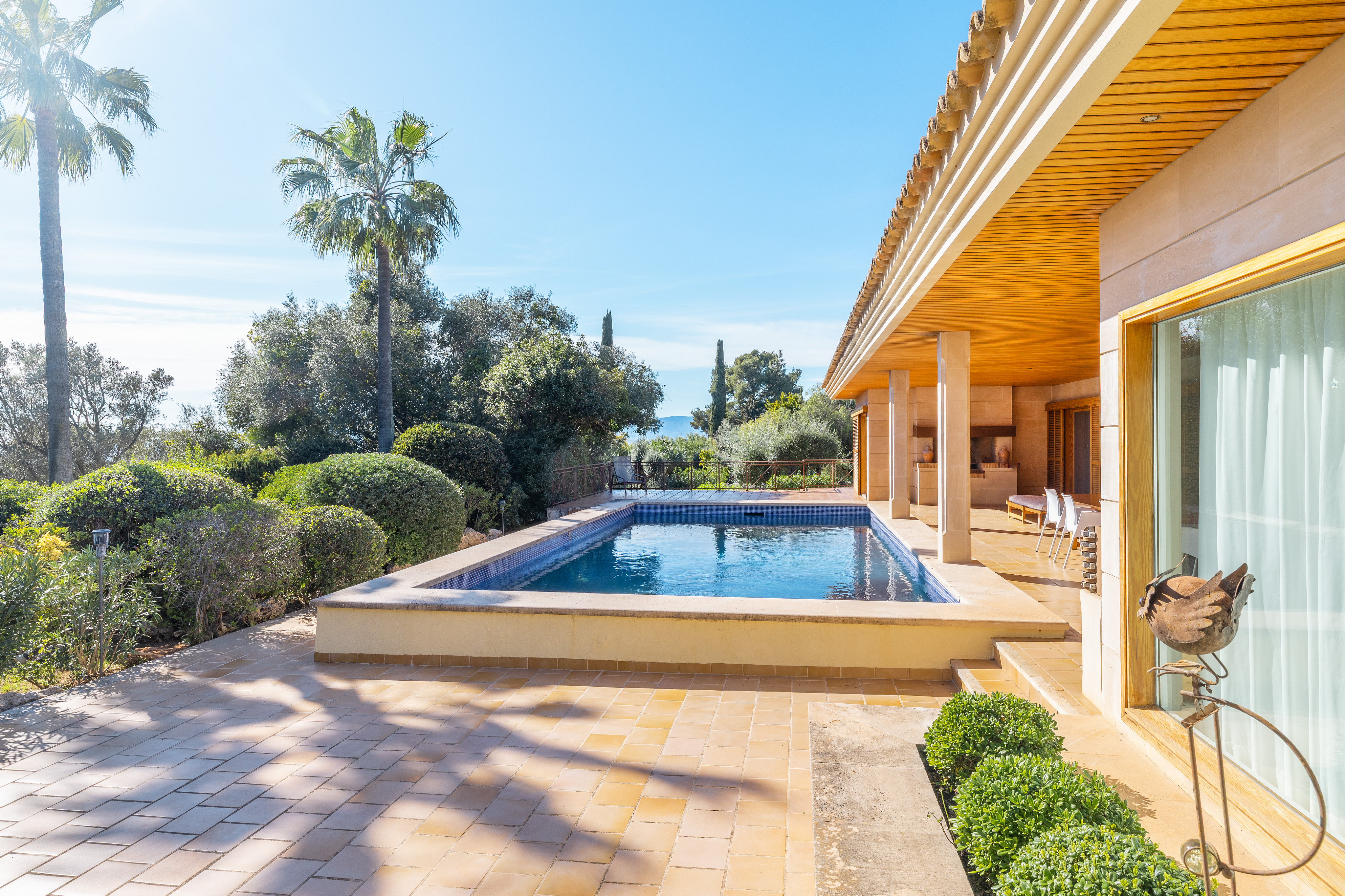 Outdoor pool with blue water, surrounded by a stone patio, lush greenery, and a tan-colored house with a wood-paneled roof.