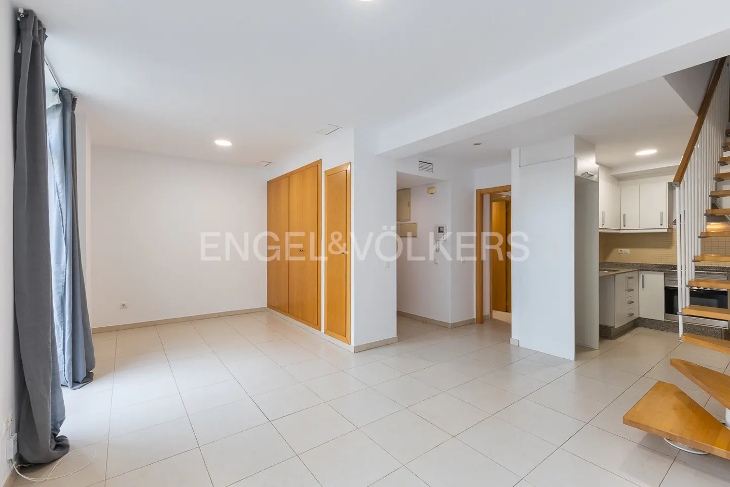 Bright, empty room with white tile floor, wood closet, and gray curtains. Open kitchen with white cabinets and wood stairs to the right.