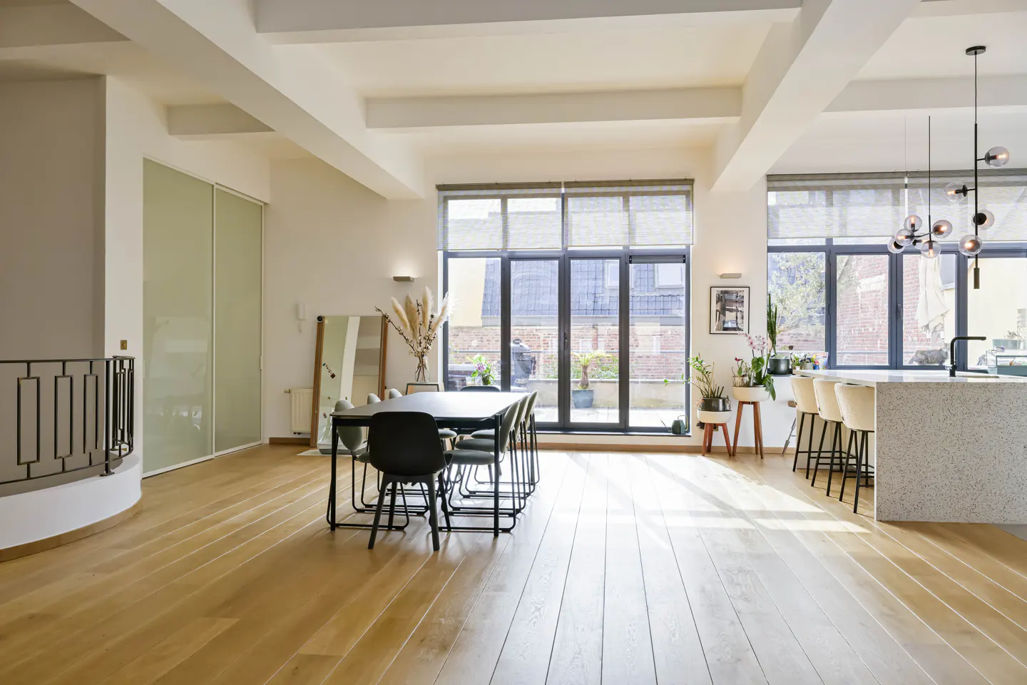 Bright, open-plan apartment with wood floors, large windows, and a dining table. A kitchen island with bar stools is visible in the background.