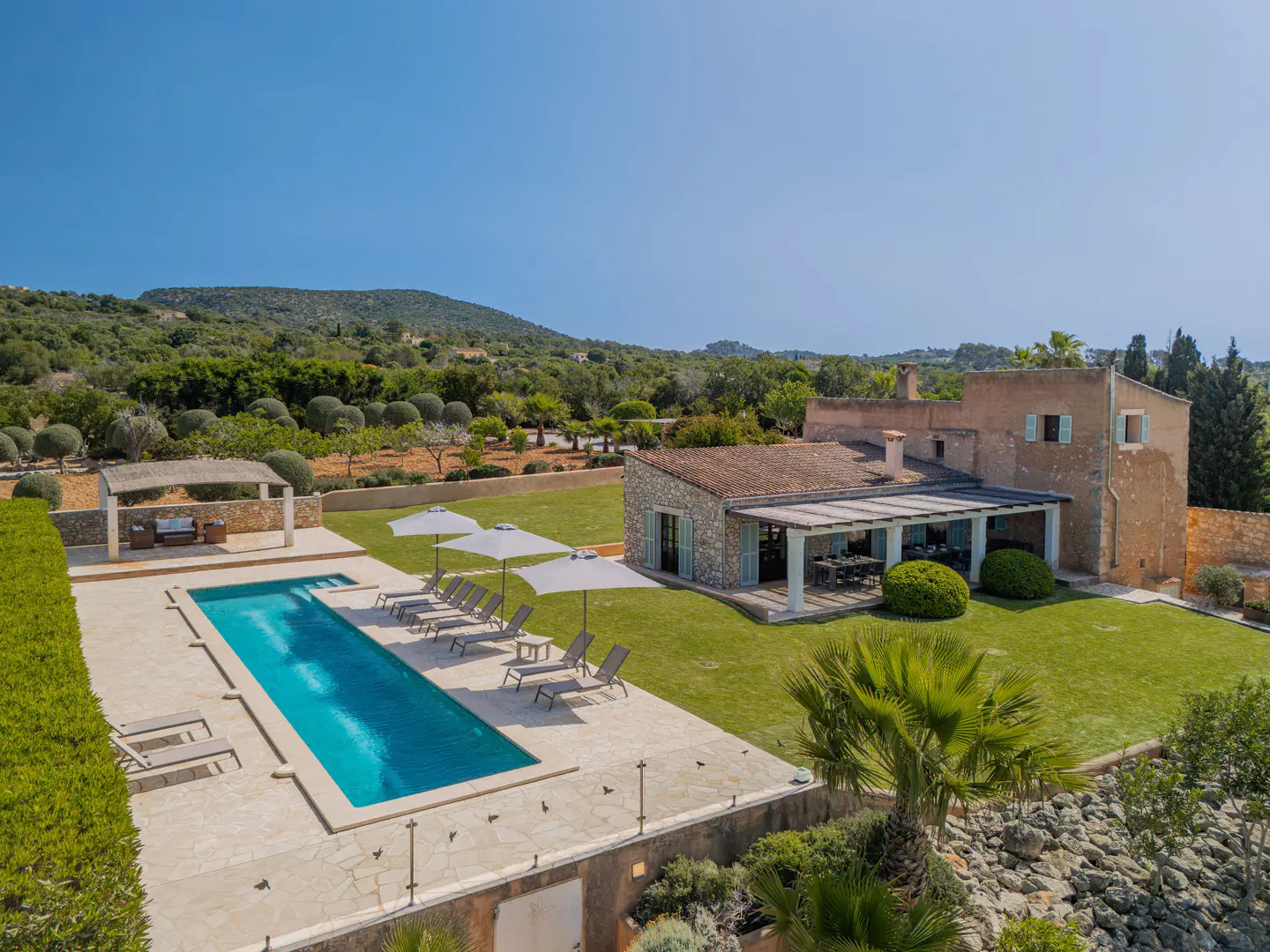Aerial view of a luxury villa with a pool, lounge chairs, and a covered patio, set against a backdrop of green hills and a clear blue sky.