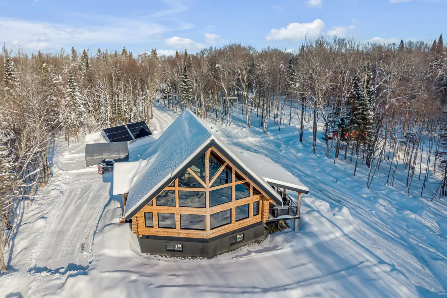 Aerial view of a snow-covered log cabin with large windows, surrounded by trees and solar panels.