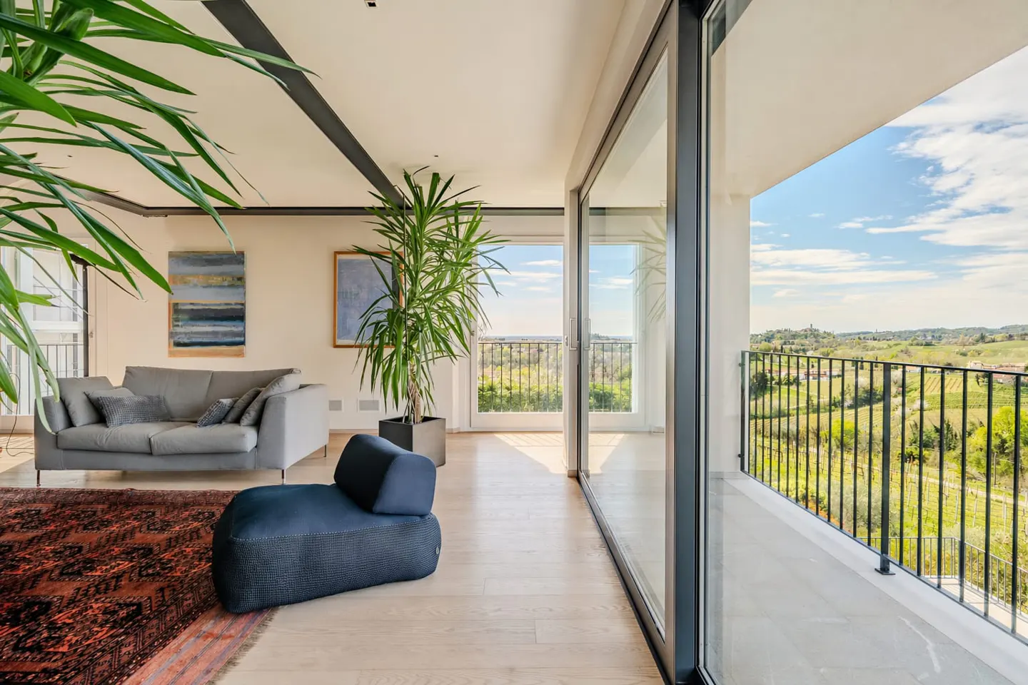 Bright living room with gray sofa, blue chair, and large windows leading to a balcony overlooking a green landscape.