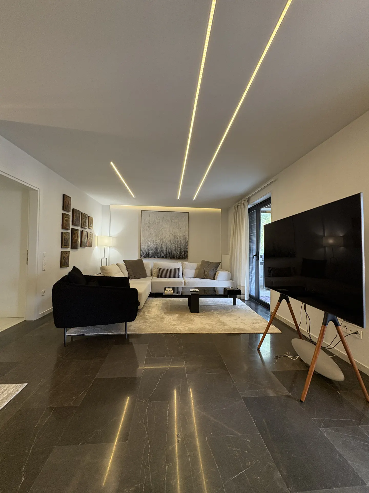 A modern living room with a black marble floor, white walls, and recessed lighting. A white sectional sofa sits on a light rug, and a large TV is on a wooden stand.