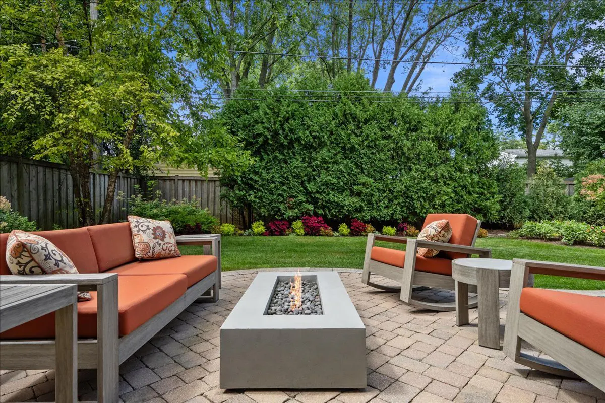 Outdoor patio with gray furniture, orange cushions, and a concrete fire pit on a brick patio. Green lawn and trees in the background.