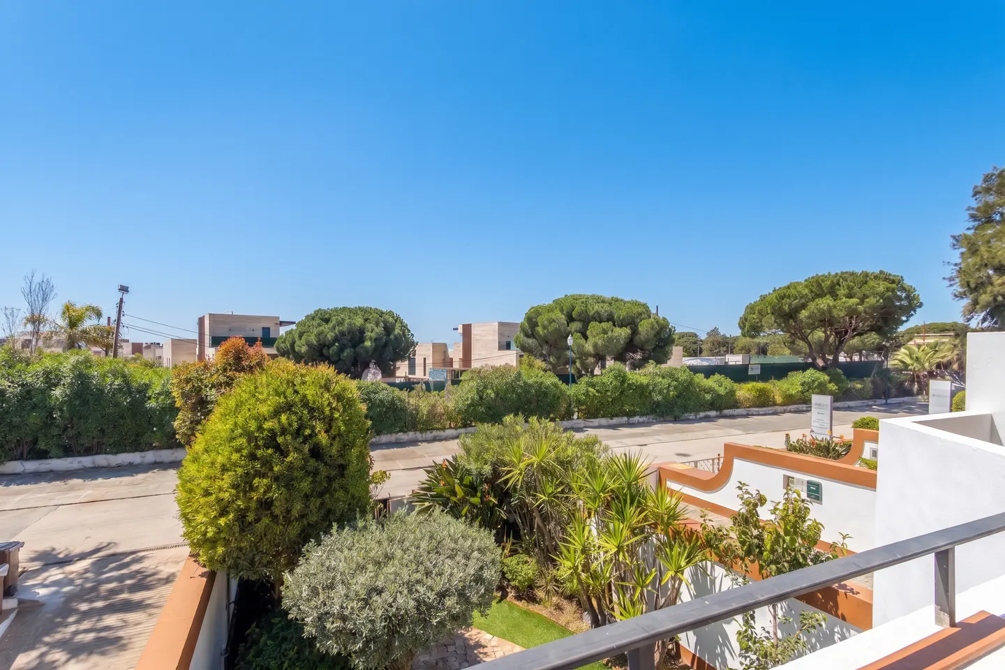 View from a balcony overlooking a garden with green bushes and trees under a clear blue sky. Buildings are visible in the background.