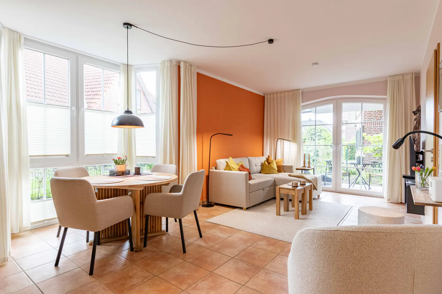 Bright living room with an orange accent wall, beige sofa, round dining table with four chairs, and large glass doors.