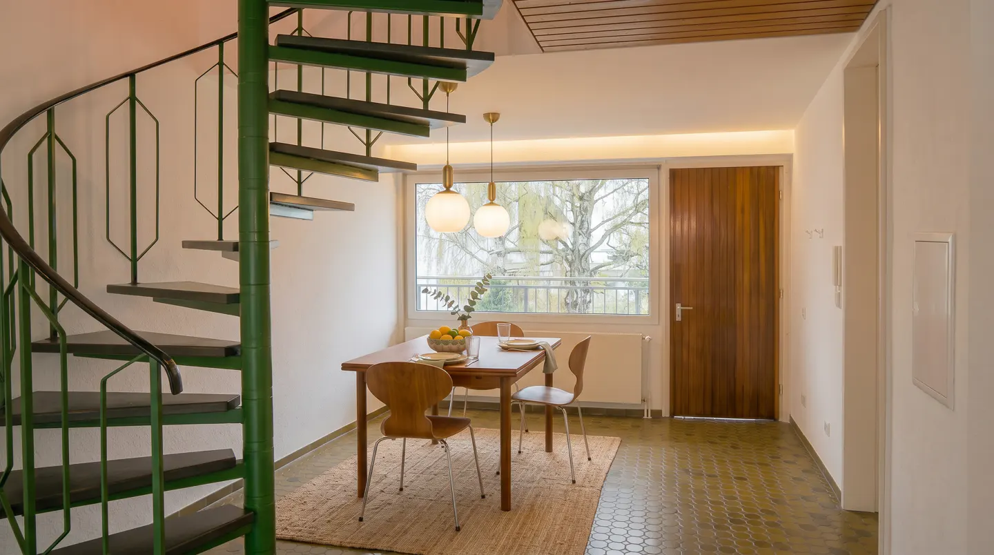Interior view of a dining area with a wooden table, chairs, and a green spiral staircase. A window overlooks a tree.