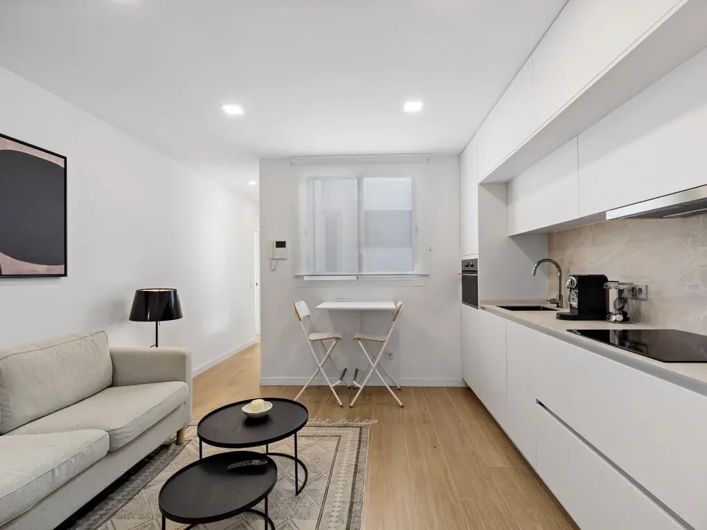 Bright, modern apartment interior with white walls, light wood floors, and a beige sofa. A black lamp and tables accent the living area, adjacent to a sleek white kitchen.