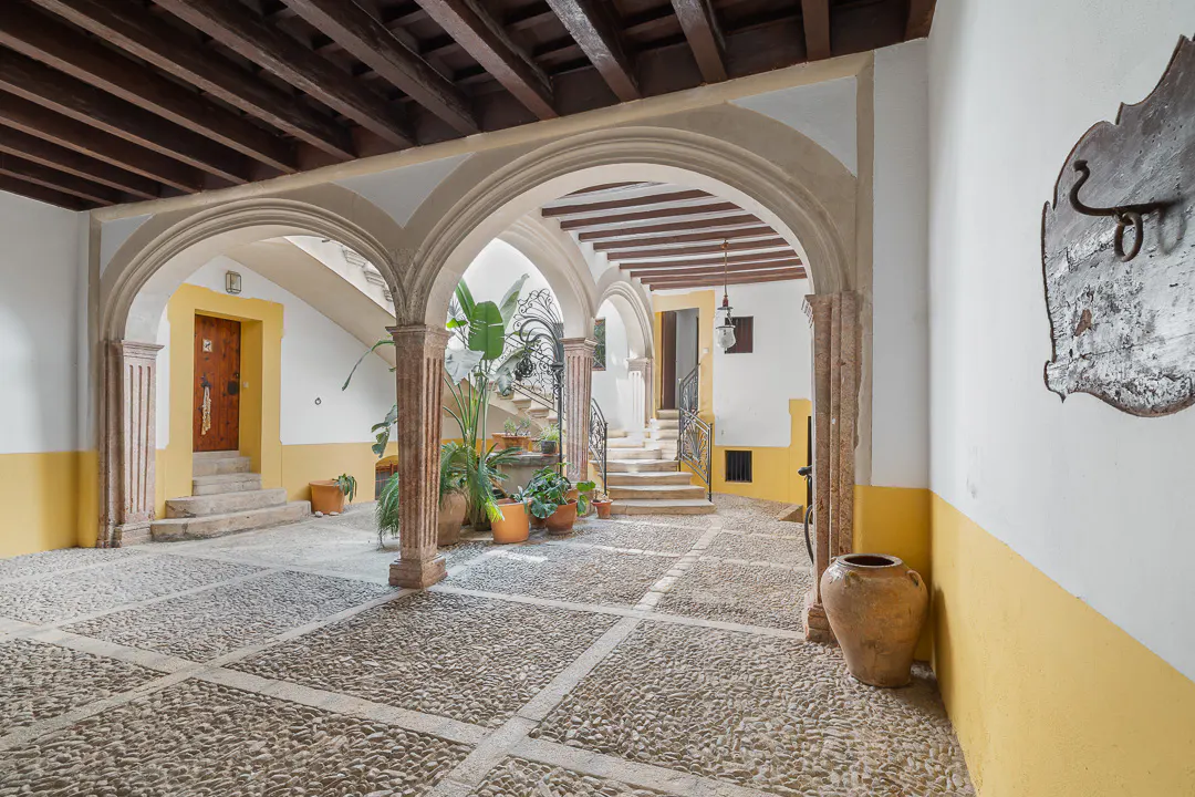 Interior courtyard with stone floors, arched walkways, and potted plants. Walls are white with yellow lower sections. Wooden beamed ceiling.