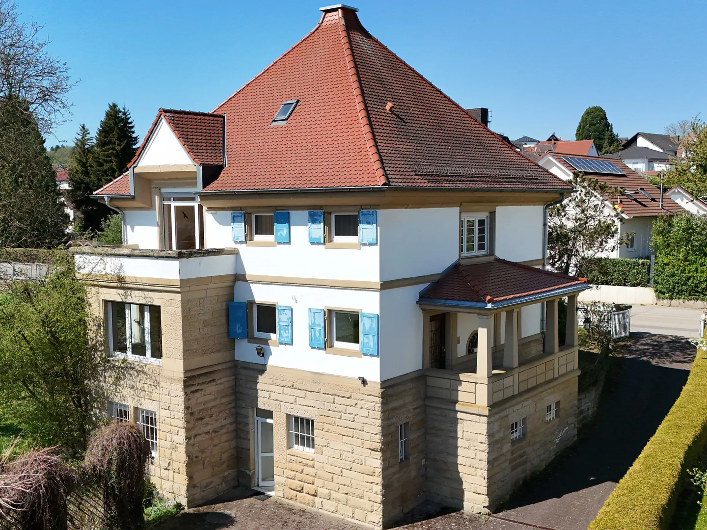 A three-story house with a red tile roof, white walls, and blue shutters. The first floor is made of stone.