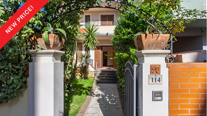 Gated entrance to a two-story house with a "New Price!" banner. Stone path leads to the open front door. Lush greenery surrounds the entrance.