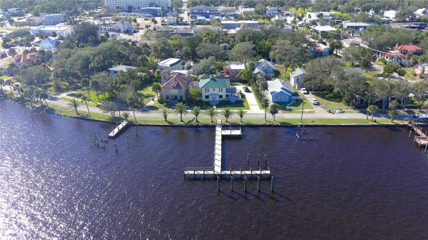 Aerial view of waterfront homes with docks on a sunny day. Houses are various colors, surrounded by trees. Dark water reflects the sunlight.