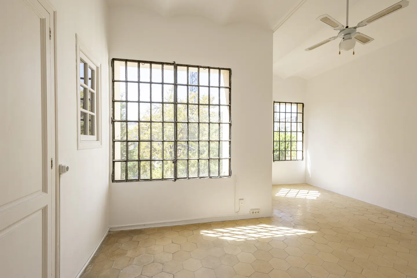 Bright, empty room with white walls, hexagon tile floor, and large windows with black frames. A ceiling fan is visible.