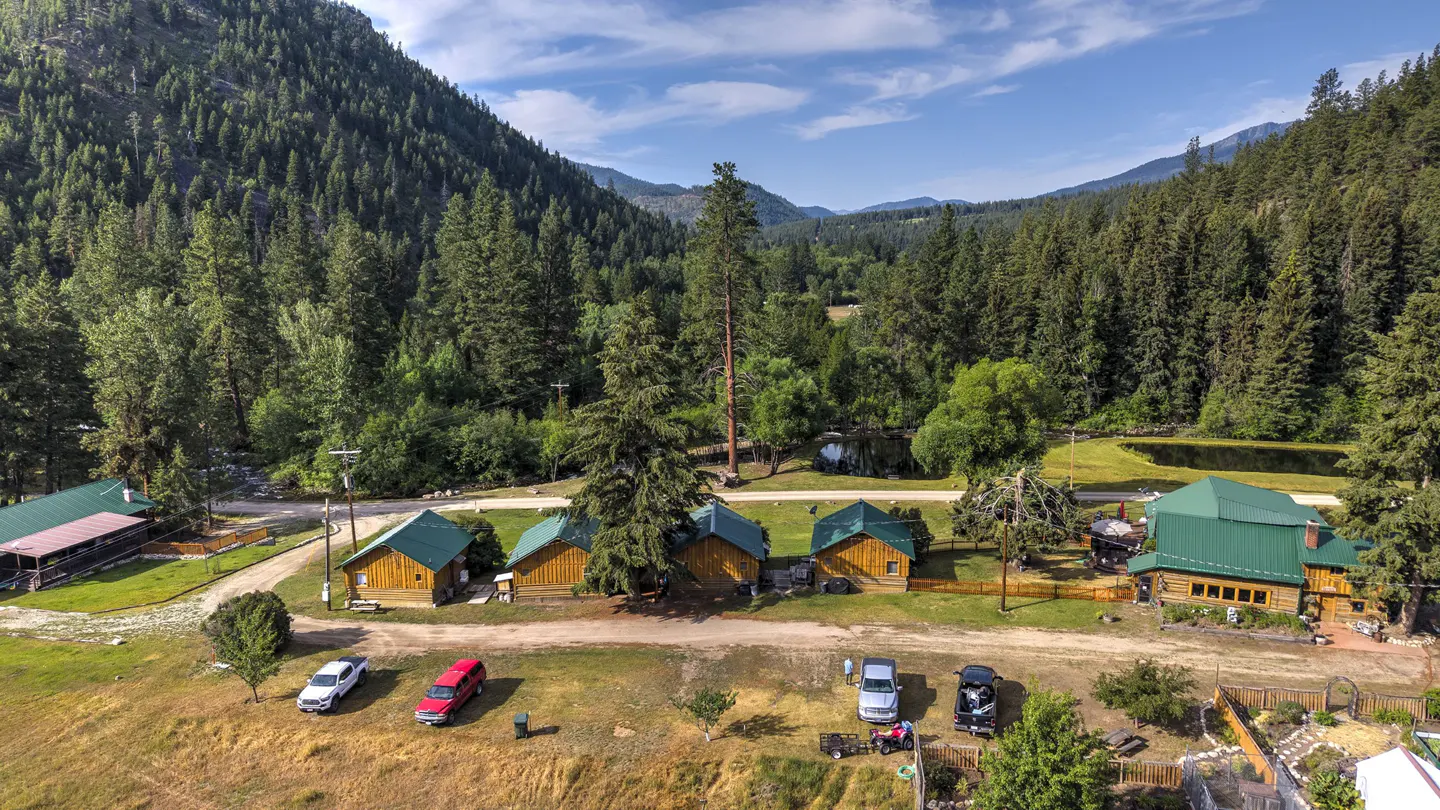 Aerial view of a wooded property with cabins and a main lodge with green roofs. Cars are parked in front. Mountains in the background.