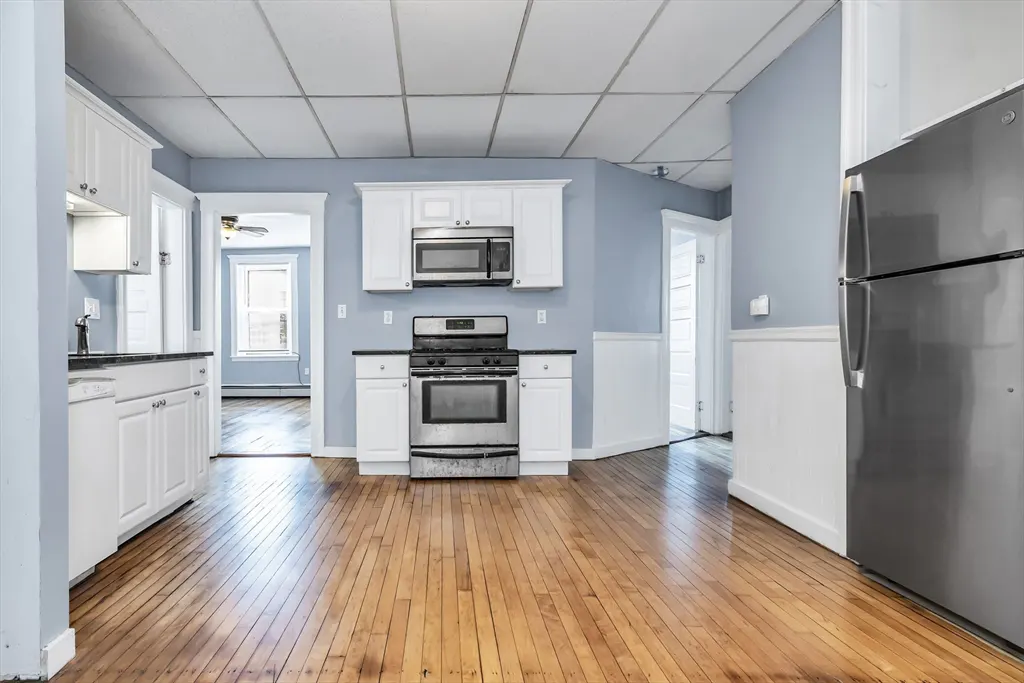 A kitchen with white cabinets, stainless steel appliances, and light blue walls. Hardwood floors and doorways to other rooms are visible.