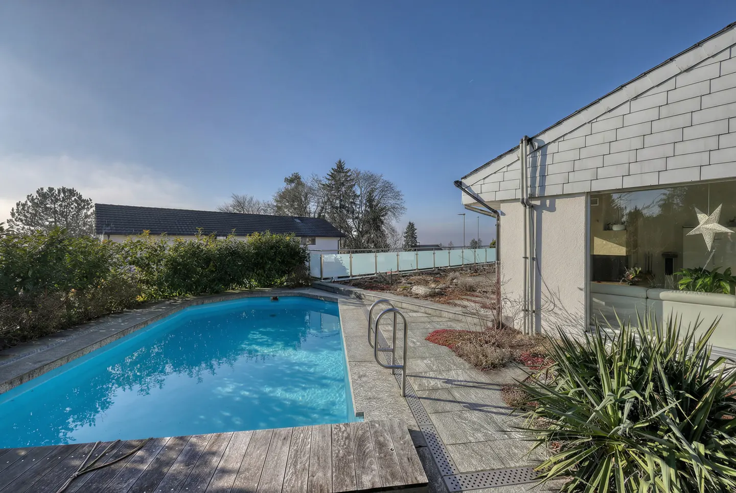 Outdoor pool with clear blue water, wooden deck, and metal ladder. A white house with a gray roof is on the right. Blue sky.