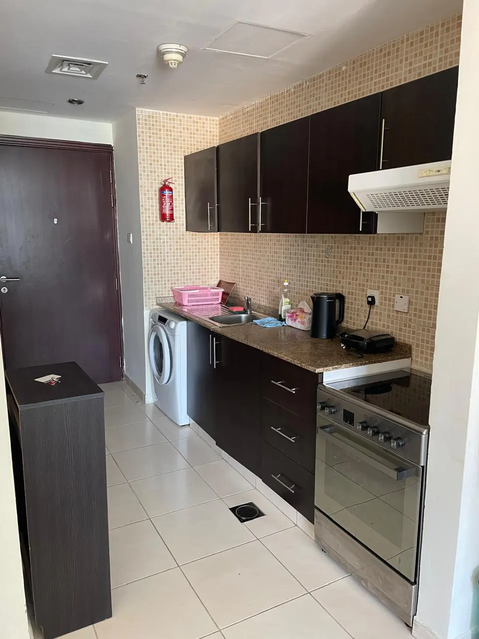 A kitchen with dark wood cabinets, beige tiled backsplash, and white tile floor. A washing machine is next to the sink. A stainless steel oven is on the right.