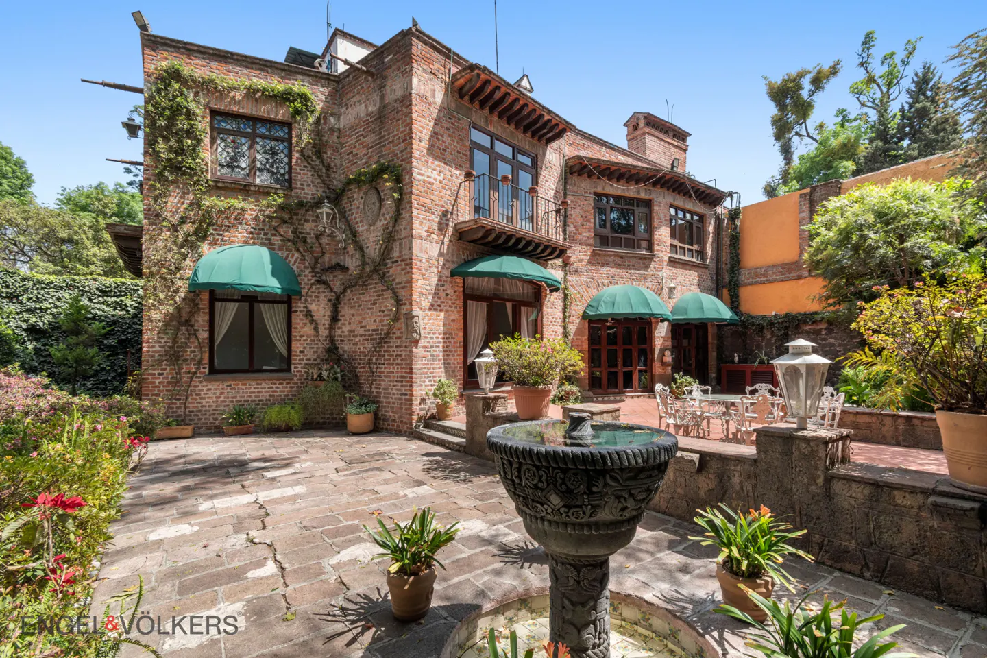 Exterior view of a brick house with green awnings, a stone patio, and a fountain.