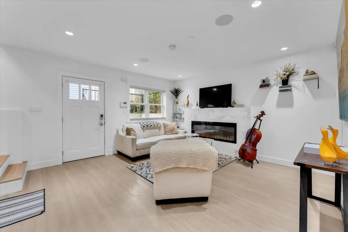 Bright living room with white walls, light wood floors, and a white sofa. A fireplace with a TV above it, and a cello leaning against the wall.