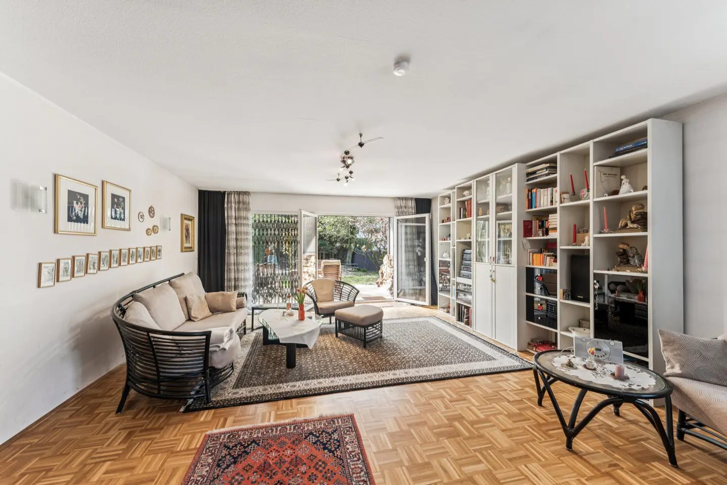 Bright living room with parquet floors, a large rug, and a white bookcase. French doors open to a garden. A curved sofa sits against a wall with framed photos.