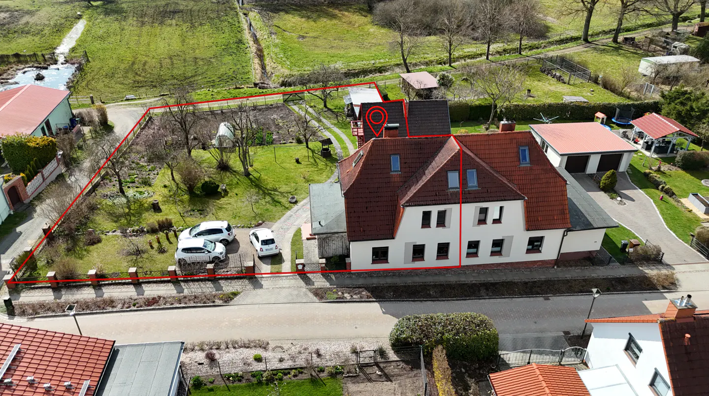 Aerial view of a two-story white house with a red roof, surrounded by a red boundary line, cars, and green space.