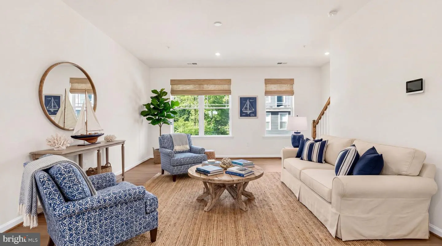 Living room with white walls, wood floors, and a jute rug. Two blue patterned armchairs, a white sofa with blue pillows, and a round wood coffee table. Coastal decor accents.