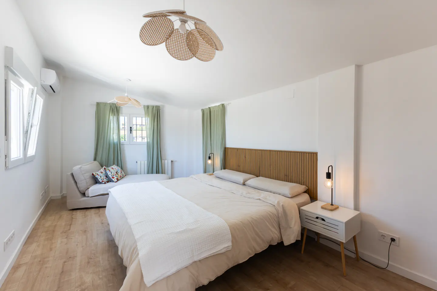 Bright bedroom with white walls, wood floors, and a large bed with a wooden headboard. A chaise lounge sits by a window with green curtains.