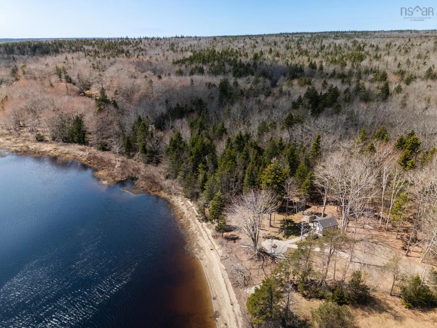 Aerial view of a lake with dark blue water, a sandy beach, and a small house nestled in the trees. Forest in the background.
