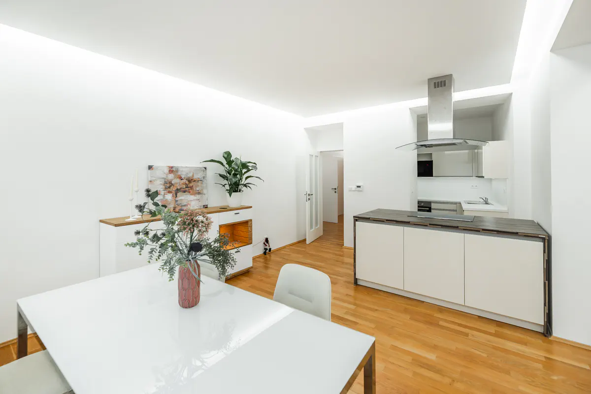 Bright, modern apartment with white walls, wood floors, and recessed lighting. A white table and chairs sit near a kitchen island with a stainless steel hood.