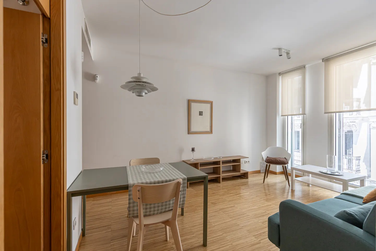 Bright living room with wood floors, white walls, and large windows. A dining table and blue sofa furnish the space.