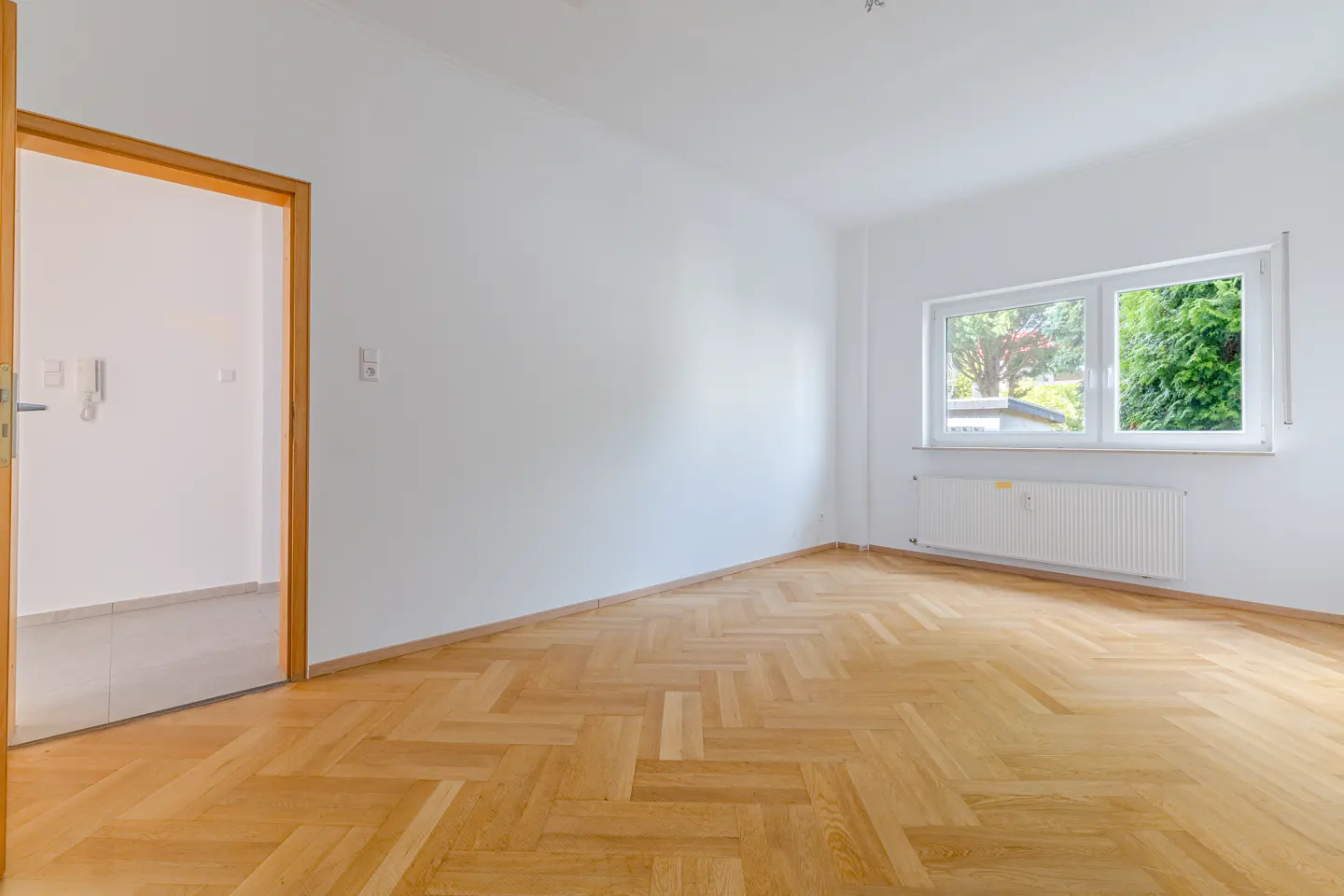 Bright, empty room with herringbone wood floor, white walls, and a window showing green trees. A wooden door frame is on the left.