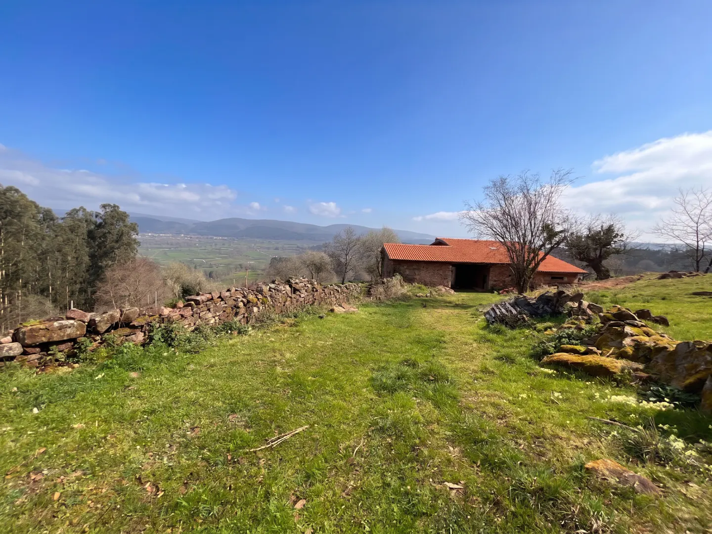 Rural property with stone house, red tile roof, and green lawn. A stone wall borders the property, with mountains in the distance under a blue sky.