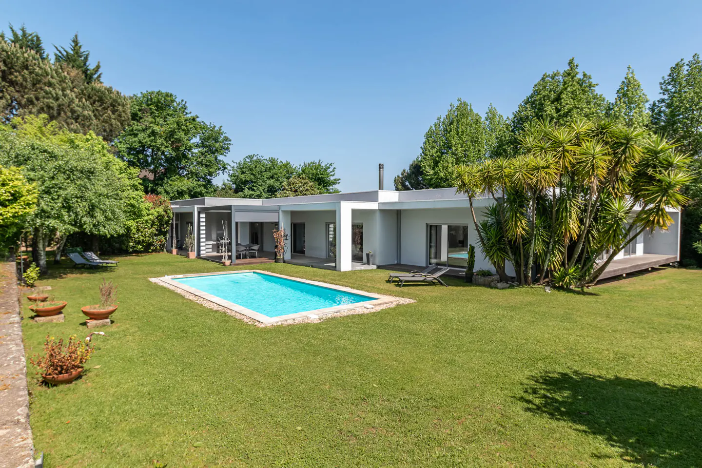 Modern white house with a blue pool and green lawn under a clear blue sky. Trees surround the property.