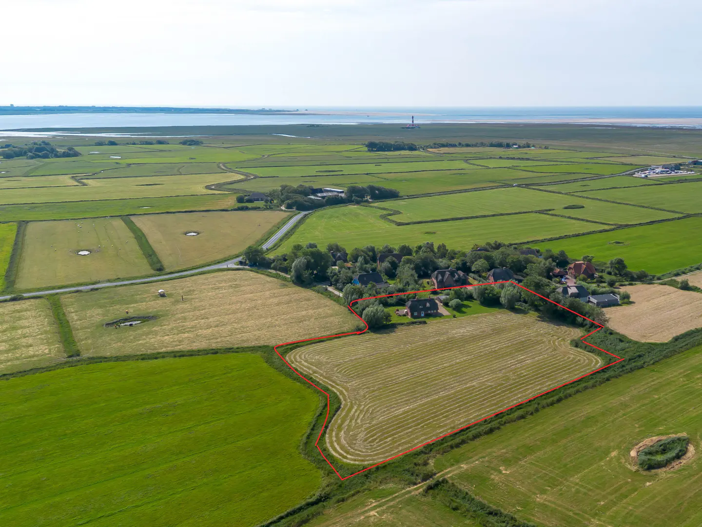 Aerial view of a rural property with a red outline, featuring green fields, houses, and a distant coastline under a bright sky.