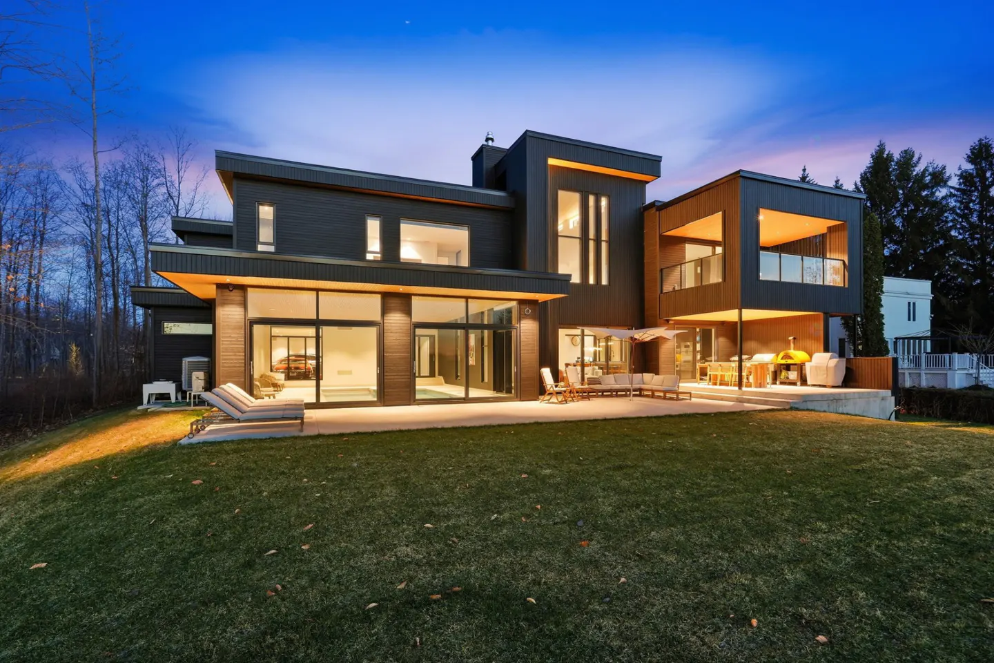 Modern dark gray house with large windows and a patio with outdoor furniture on a green lawn at dusk.