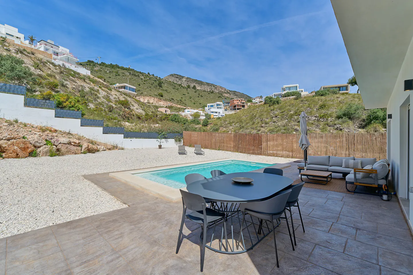 Outdoor patio with a pool, table, and seating area. Hillside homes are visible in the background.