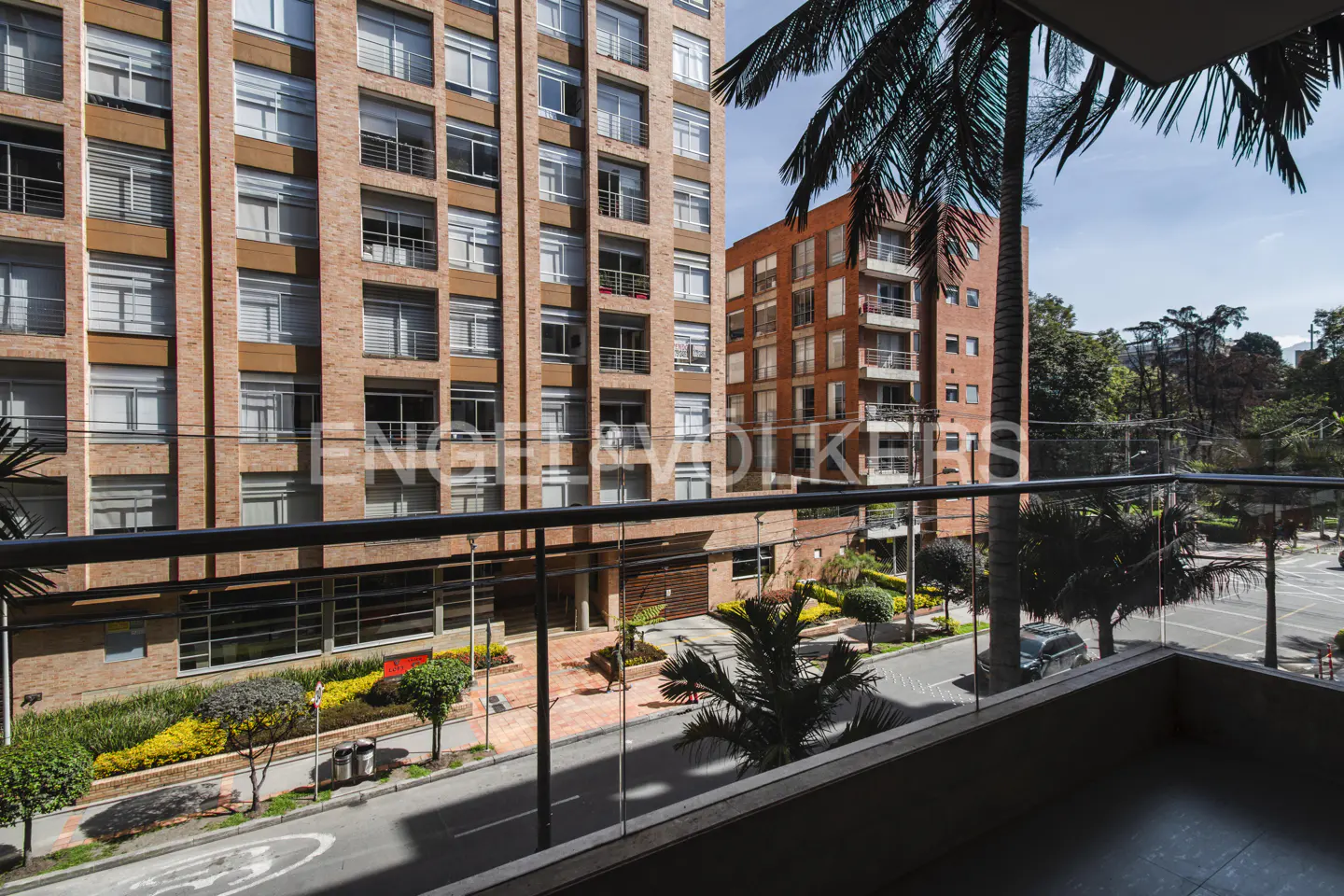 View from a balcony with glass railings, overlooking a street with brick buildings and trees.