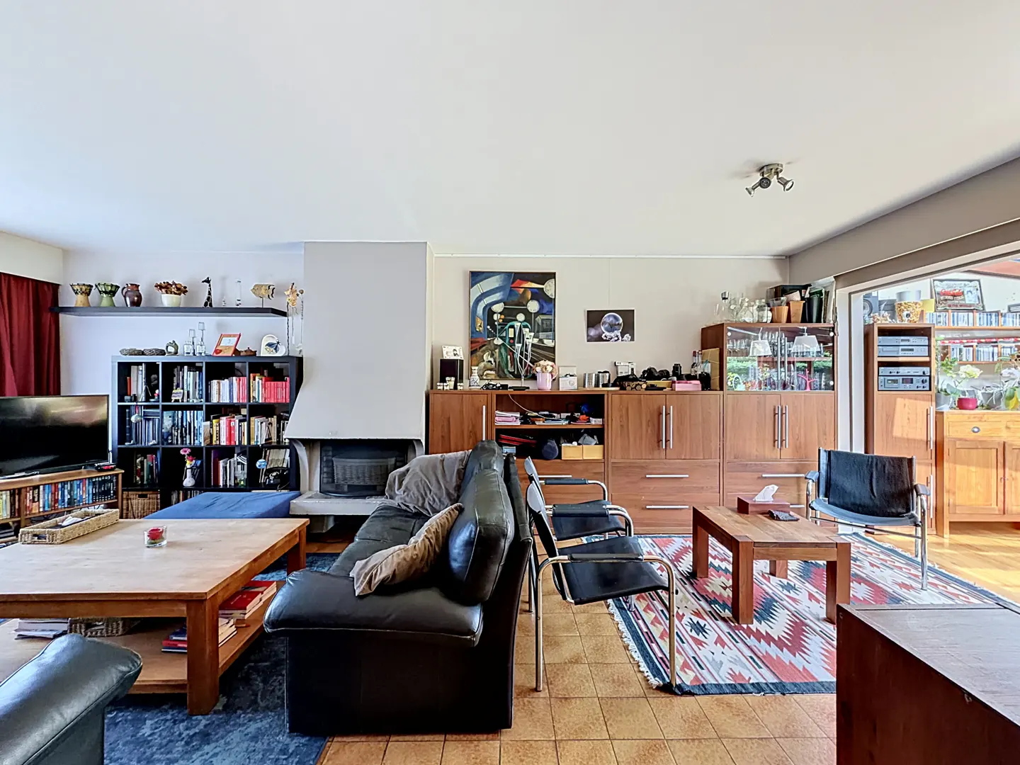Living room with a black leather sofa, fireplace, bookshelves, and wooden furniture. A patterned rug sits on the tiled floor.