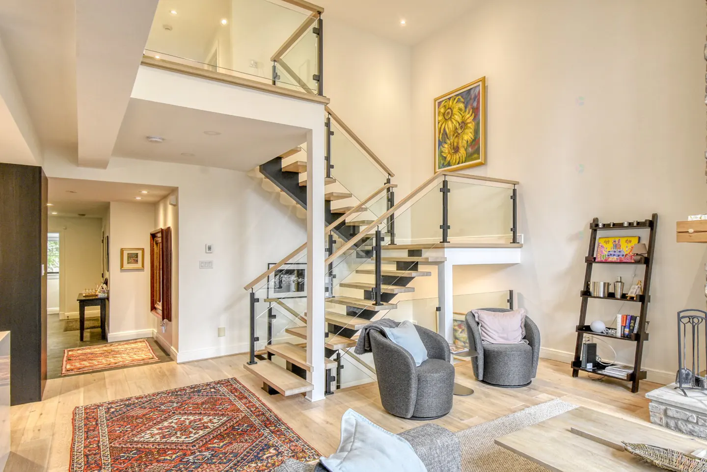 Bright living room with hardwood floors, a red patterned rug, and a modern staircase with glass railings. Two gray chairs face the stairs.
