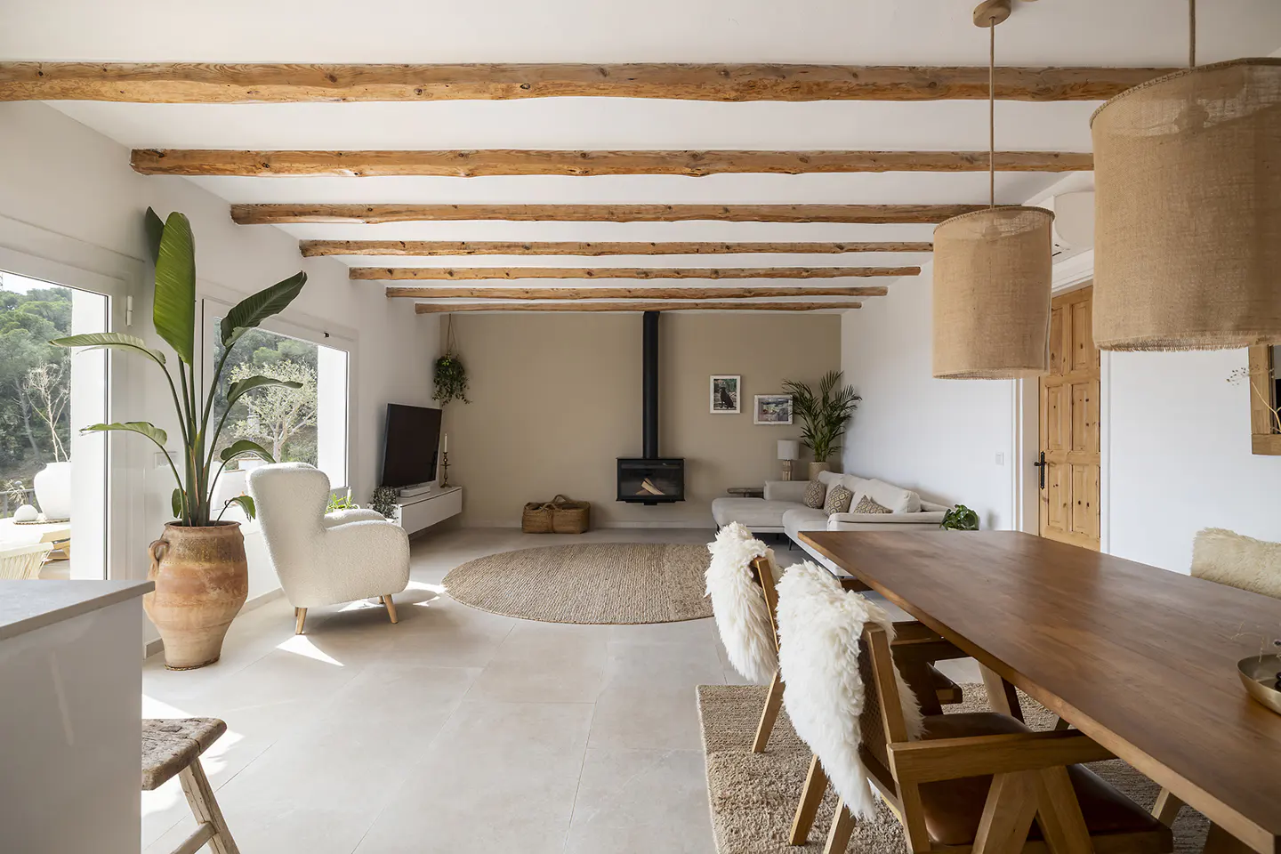Bright living room with wooden beams, a fireplace, and a large wooden table with chairs. A large plant sits near a window.
